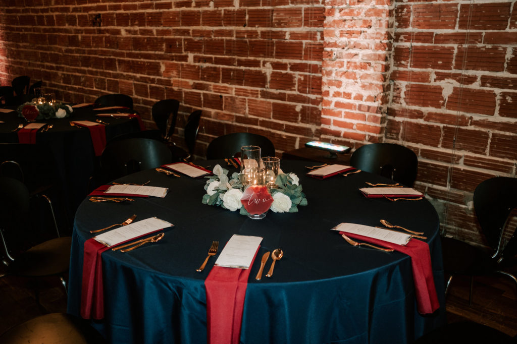 Industrial Florida Wedding Reception, Dark and Moody Decor against Exposed Brick Wedding Venue Round Tables with Dark Navy Linens, Burgundy Napkins and Gold Flatware, Low Faux Floral Centerpieces with Floating Candle Centerpieces | Downtown St. Pete Wedding Venue NOVA 535
