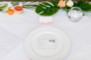 Tropical Florida Citrus Wedding Centerpieces with Cut Oranges Lemons and Grapefruit and Monstera Palm Greenery Garland Runner | Wedding Reception Place Setting with Pink Napkin, Pink Place Card, and Postcard Favor
