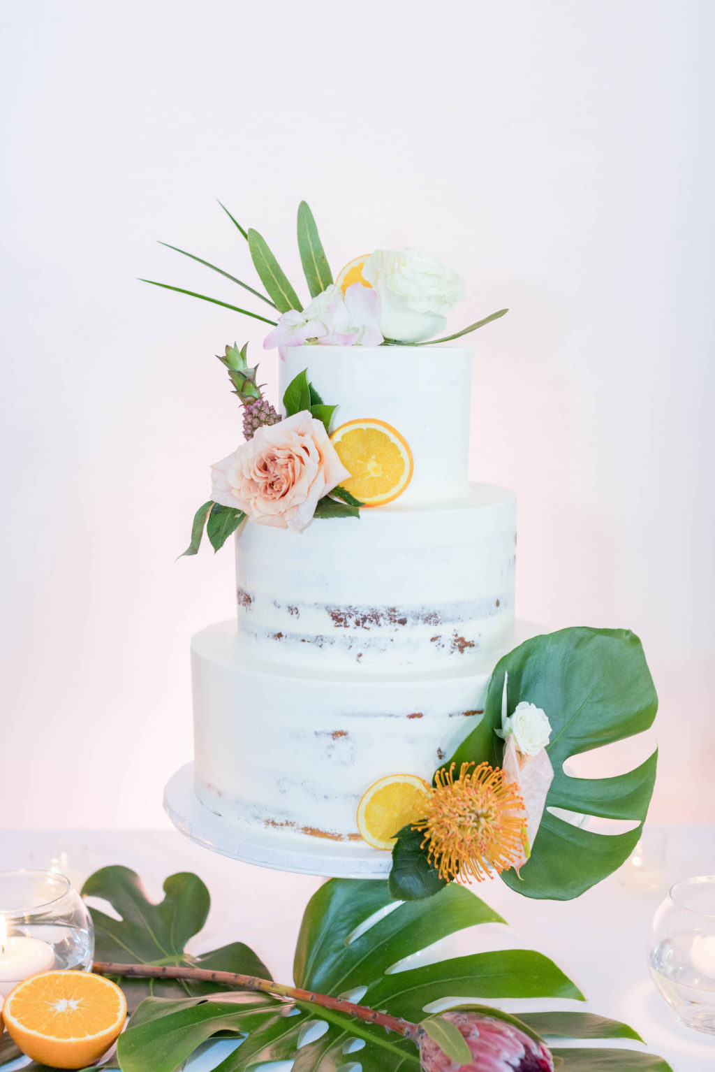 Tropical Beach Wedding Cake | Three Tier Semi Naked Half Iced Buttercream Wedding Cake with Tropical Greenery Monstera Leaf, Orange Slices and Mini Ornamental Pineapple, Blush Pink Roses and Protea by The Artistic Whisk
