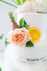 Tropical Beach Wedding Cake | Three Tier Semi Naked Half Iced Buttercream Wedding Cake with Tropical Greenery Monstera Leaf, Orange Slices and Mini Ornamental Pineapple, Blush Pink Roses and Protea by The Artistic Whisk