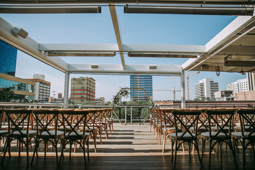 Outdoor Rooftop Downtown St. Petersburg Wedding Ceremony at St. Pete Wedding Venue Red Mesa Events | Wood Cross Back French Country Chairs | Geometric Gold Arch Ceremony Backdrop with Natural Rustic Floral Spray of White Roses and Eucalyptus Greenery | Perfecting the Plan