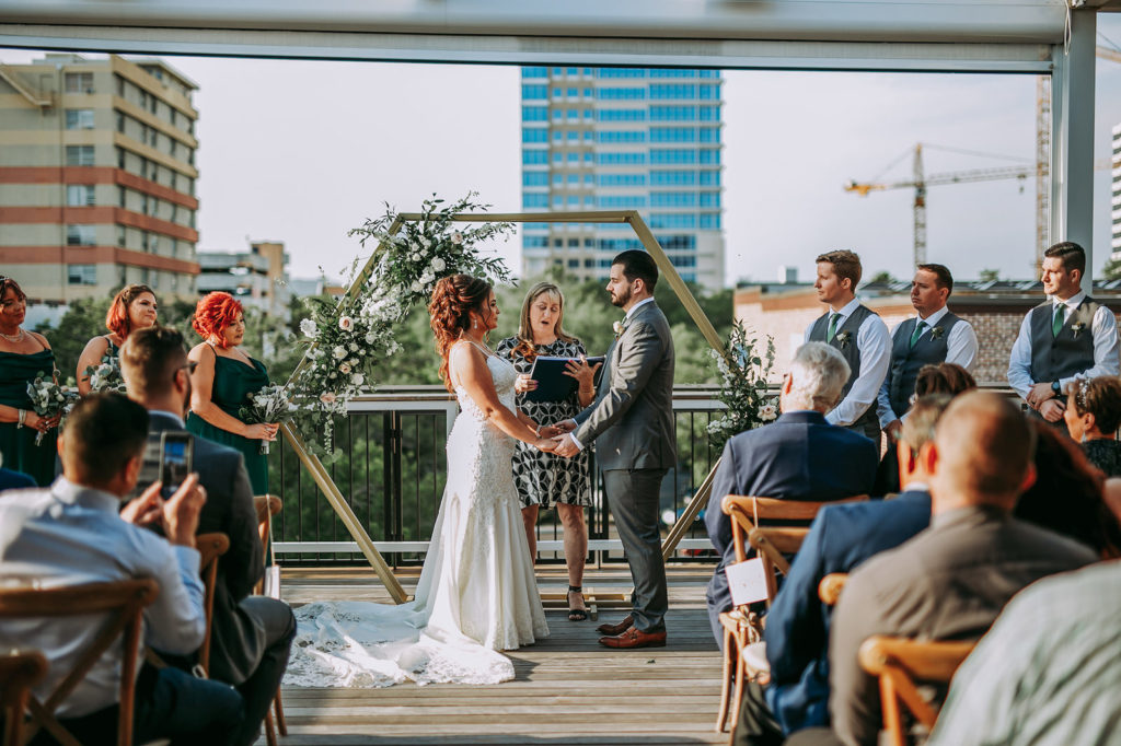 Bride and Groom Exchanging Vows During Outdoor Rooftop Downtown St. Petersburg Wedding Ceremony at St. Pete Wedding Venue Red Mesa Events | Wood Cross Back French Country Chairs | Geometric Gold Arch Ceremony Backdrop with Natural Rustic Floral Spray of White Roses and Eucalyptus Greenery | Perfecting the Plan