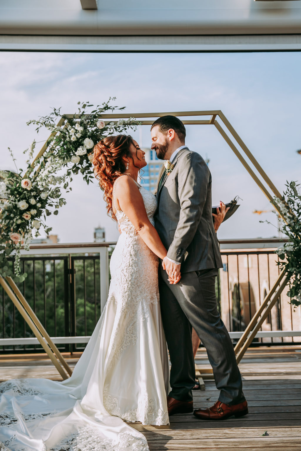 Bride and Groom Exchanging Vows During Outdoor Rooftop Downtown St. Petersburg Wedding Ceremony | Lace Mermaid Wedding Dress | Charcoal Grey Groom Suit | Geometric Gold Arch Ceremony Backdrop with Natural Rustic Floral Spray of White Roses and Eucalyptus Greenery