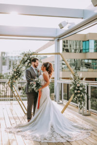 Outdoor Rooftop Bride and Groom Portrait in front of Geometric Gold Arch Ceremony Backdrop with Natural Rustic Floral Spray of White Roses and Eucalyptus Greenery | Groom Wearing Classic Charcoal Grey Suit with Green Tie | Lace Fit and Flare Mermaid Spaghetti Strap Wedding Dress Bridal Gown with Scallop Edge Lace Train Hem