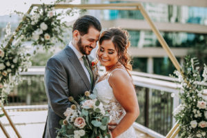 Outdoor Rooftop Bride and Groom Portrait in front of Geometric Gold Arch Ceremony Backdrop with Natural Rustic Floral Spray of White Roses and Eucalyptus Greenery | Groom Wearing Classic Charcoal Grey Suit with Green Tie | Perfecting the Plan