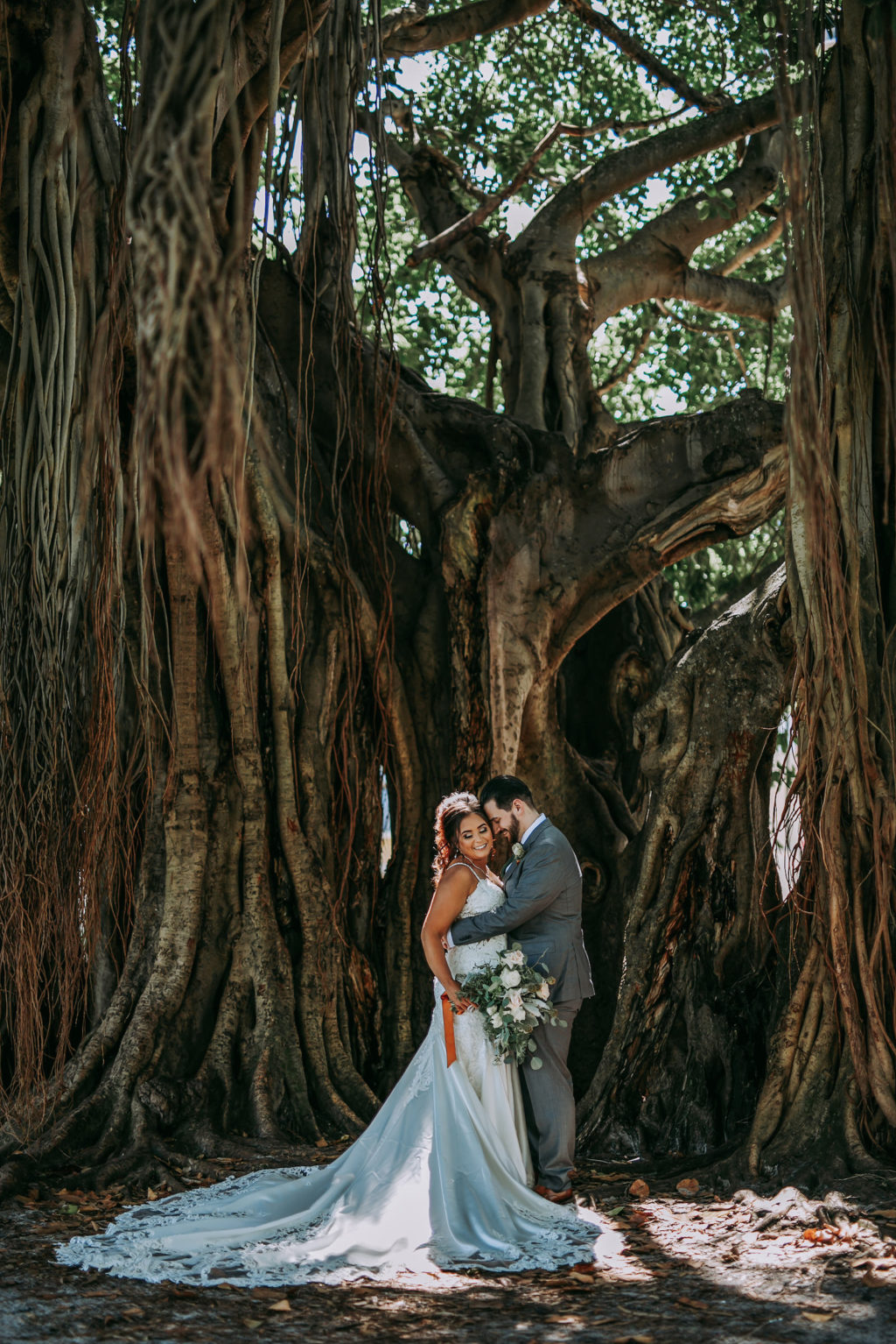 Outdoor Bride and Groom Portrait in front of Banyan Trees in Vinoy Park Downtown St. Pete Florida | Groom Wearing Classic Charcoal Grey Suit with Green Tie | Lace Fit and Flare Mermaid Spaghetti Strap Wedding Dress Bridal Gown with Scallop Edge Lace Train Hem