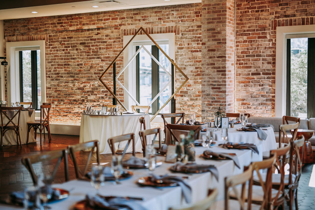 Indoor Historic Venue Brick Wall Wedding Reception at Downtown St. Pete Wedding Venue Red Mesa Events | Cross Back Wood French Country Chairs with White Table Linens on Long Feasting Tables | Gold Geometric Arch Sweetheart Table Backdrop | Perfecting the Plan