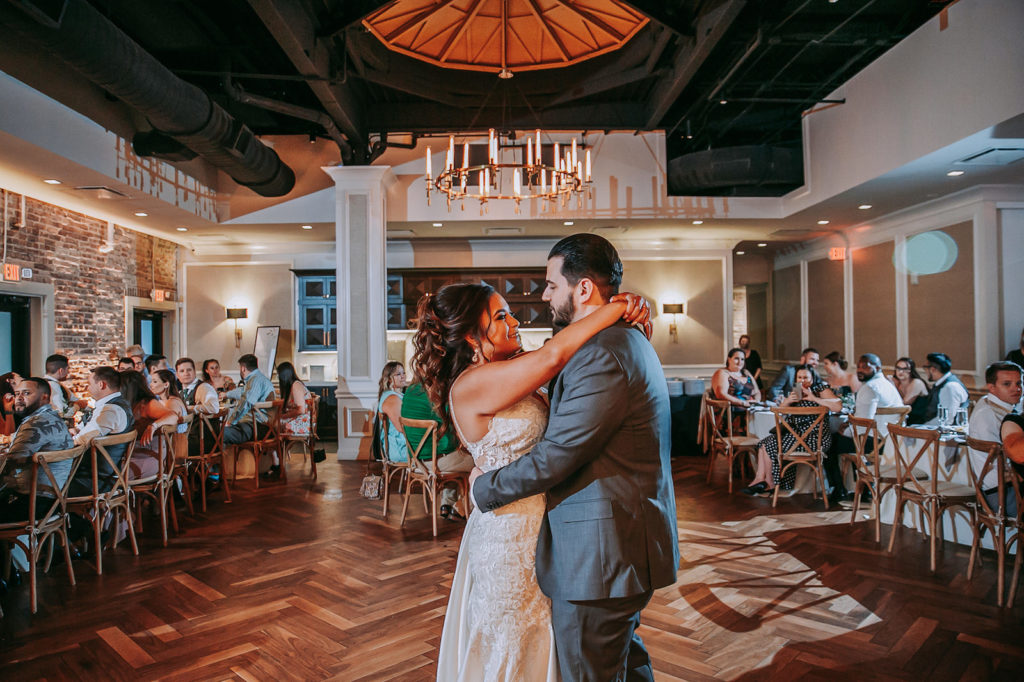 Bride and Groom First Dance at Indoor Historic Venue Brick Wall Wedding Reception at Downtown St. Pete Wedding Venue Red Mesa Events | Cross Back Wood French Country Chairs with White Table Linens on Long Feasting Tables