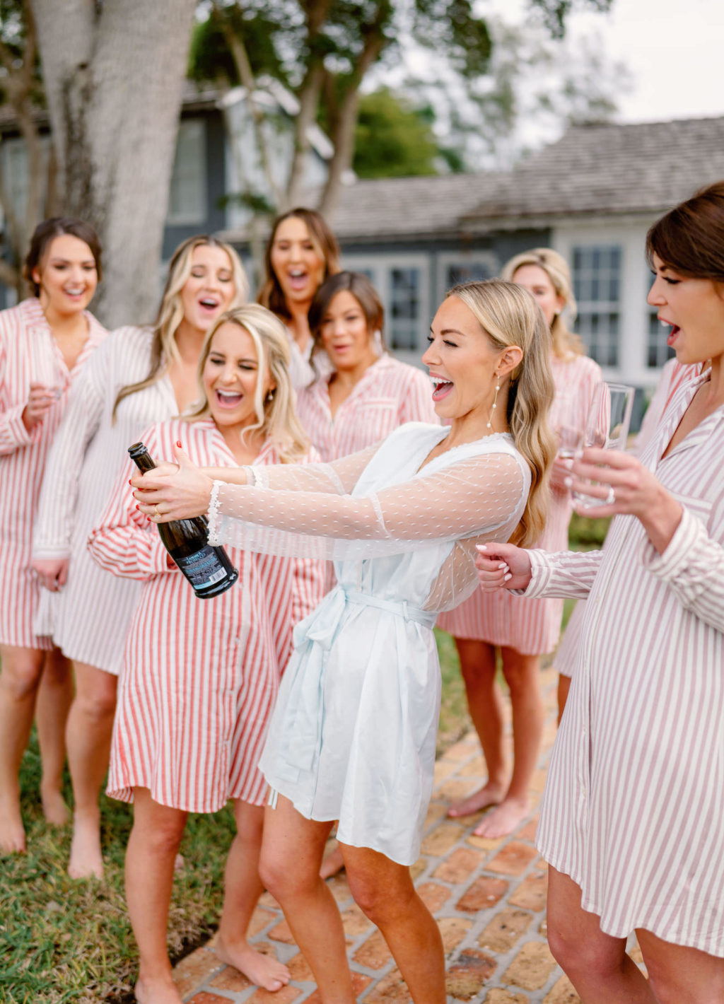 Elegant Bride in Light Blue with Illusion Sleeves Robe Getting Wedding Ready with Bridesmaids in Pinstripe Pajamas Popping Bottle of Champagne