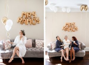 Florida Bride Getting Wedding Ready in White Robe Sitting on Couch Holding Gold and White Latex Balloons, Letter Foil Balloons Backdrop with New Last Name, Bridesmaids in Blue Robes Popping Bottle of Champagne | Tampa Bay Wedding Photographer Lifelong Photography Studio