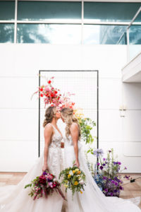 Gay LGBTQ+ Pride Wedding, Lesbian Brides Holding Hands with Red and Yellow Floral Bouquets, Black Metal Grid Mesh Rectangle Arch with Rainbow Flower Arrangement, Pink, Orange, Purple and Blue | Tampa Bay Wedding Planner Stephany Perry Events | Rooftop Wedding Venue Hotel Alba Tampa