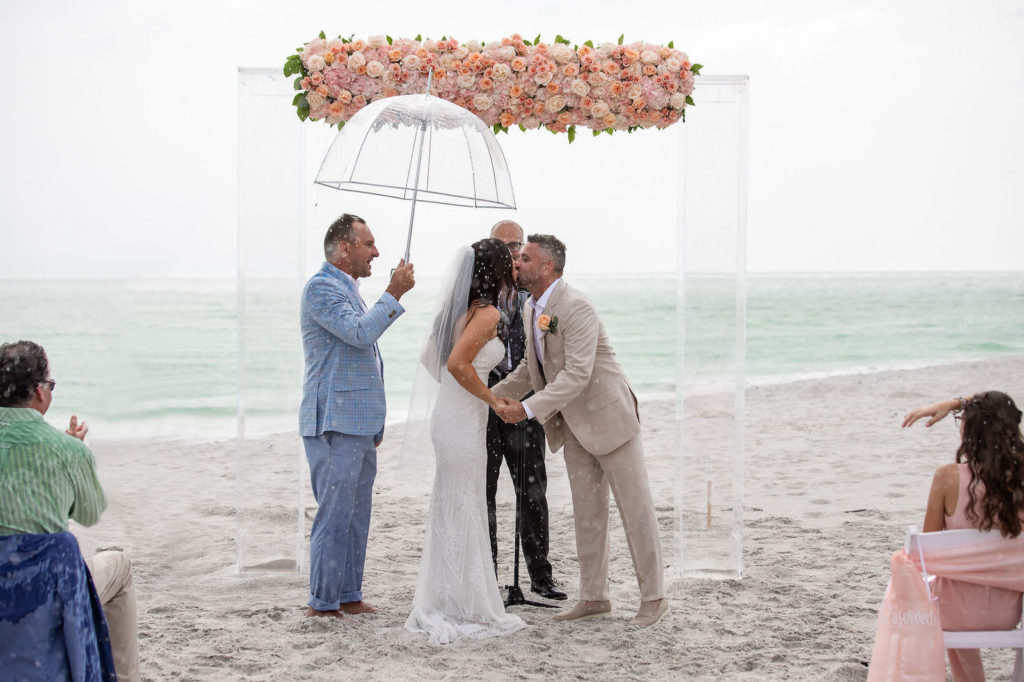 Wedding Day Rain Storm with Clear Umbrella | Bride and Groom First Kiss at Florida Beach Wedding at Sarasota Wedding Venue The Resort at Longboat Key Club | Ceremony Backdrop with Clear Acrylic Arch and Floral Arrangement of Peach Pink and Coral Roses | Bride Wearing Strapless Sheath Wedding Gown by Tara Keilly | Groom Wearing Casual Khaki Suit