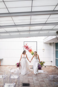 Gay LGBTQ+ Pride Wedding, Lesbian Brides Holding Hands with Red and Yellow Floral Bouquets, Black Metal Grid Mesh Rectangle Arch with Rainbow Flower Arrangement, Pink, Orange, Purple and Blue | Tampa Bay Wedding Planner Stephany Perry Events | Rooftop Wedding Venue Hotel Alba Tampa