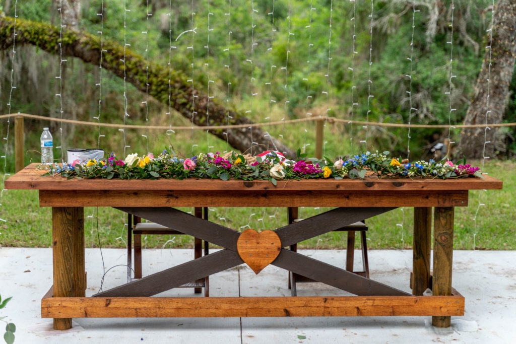 Intimate Backyard DIY Tampa Wedding Reception Decor, Wooden Table with Heart, Greenery Garland with Purple, Pink and Blue Flowers, Hanging String Lights