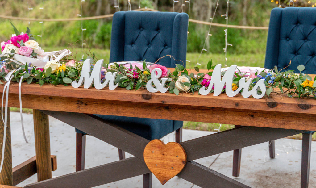 Intimate Backyard DIY Tampa Wedding Reception Decor, Wooden Table with Heart, Greenery Garland with Purple, Pink and Blue Flowers, Hanging String Lights , Cut Out Mr & Mrs Sign, Navy Blue Tufted Groom and Bride Seats