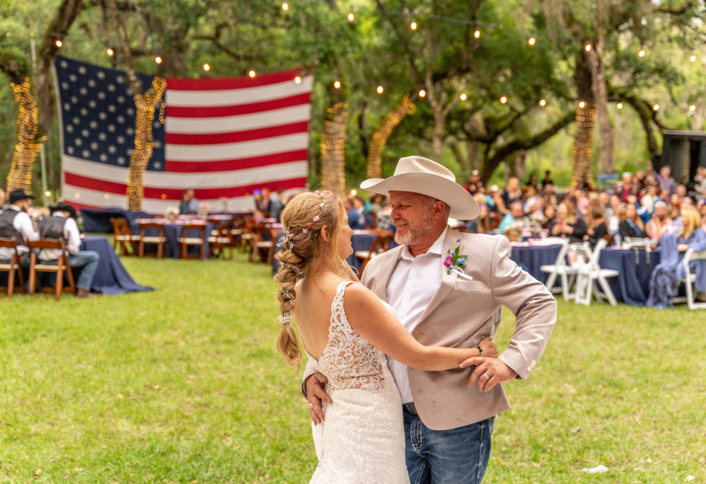 Backyard Intimate DIY Tampa Bride in Lace Open Back Wedding Dress, Braided Hair with Flowers, Groom in Tan Suit Jacket and Jeans, Cowboy Hat First Dance Wedding Reception, American Flag Hanging in Backdrop with String Lights