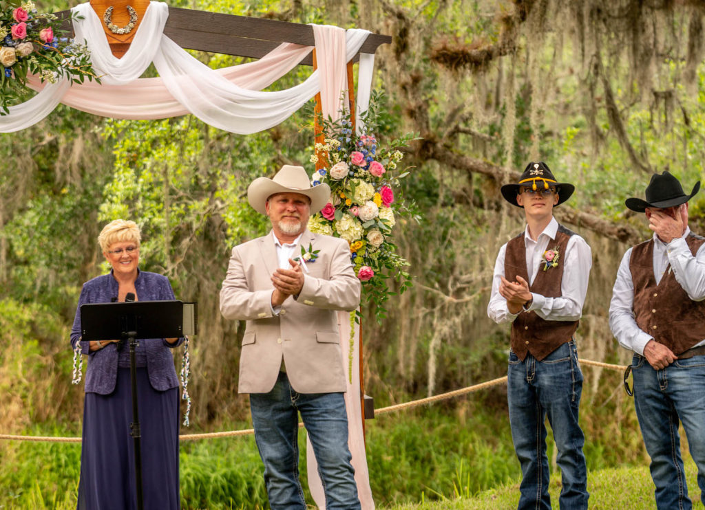 Tampa Intimate DIY Backyard Wedding Ceremony, Groom in Jeans and Tan Suit Jacket, Cowboy Hat Watching Emotional Reaction to Bride, Wooden Arch with Blush Pink and White Linen Draping, Colorful Pink, White and Blue with Greenery Floral Arrangements, Groomsmen in Brown Vests and Black Cowboy Hats