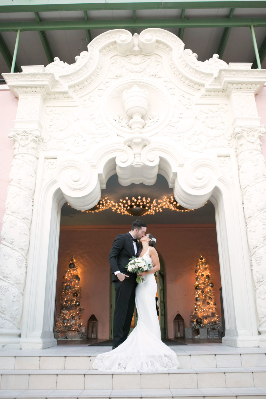 Elegant Classic Bride in Romantic Lace Train and Crepe Fitted Wedding Dress Holding White and Greenery Floral Bouquet, Groom in Classic Black Tux Under Arch of St. Pete Wedding Venue The Vinoy Renaissance | Tampa Bay Wedding Photographer Carrie Wildes Photography | Wedding Dress Shop Isabel O'Neil Bridal Collection