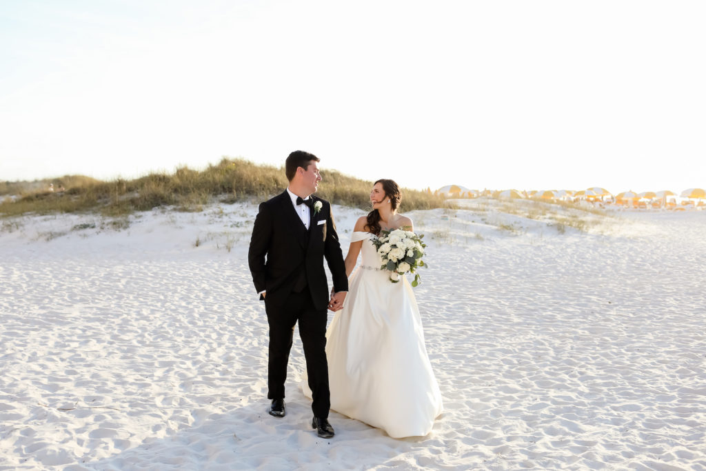 Timeless Florida Bride Wearing Off the Shoulder A-Line Wedding Dress with Rhinestone Belt Holding White Roses and Greenery Floral Bouquet and Groom in Classic Black Tuxedo on White Sand Beaches | Tampa Bay Wedding Photographer Lifelong Photography Studio | Clearwater Beach Wedding Venue Sandpearl Resort | Wedding Florist Iza's Flowers | Wedding Hair and Makeup Adore Bridal