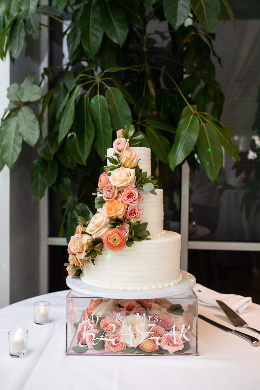 Three Tier Buttercream Striped Icing Wedding Cake with Fresh Flowers of Peach Roses and Coral Ranunculus and Greenery Cascading down the Side | Square Clear Acrylic Cake Stand filled with Peach and Pink Fresh Flowers Roses with Custom Bride and Groom Name Decal