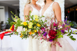 Colorful Modern Rainbow Gay LGBTQ+ Pride Wedding Reception Decor, Lesbian Brides Holding Yellow, Pink and Purple Wild Floral Bouquets, White Flower Wall Backdrop | Tampa Bay Wedding Planner Stephany Perry Events | Wedding Venue Hotel Alba