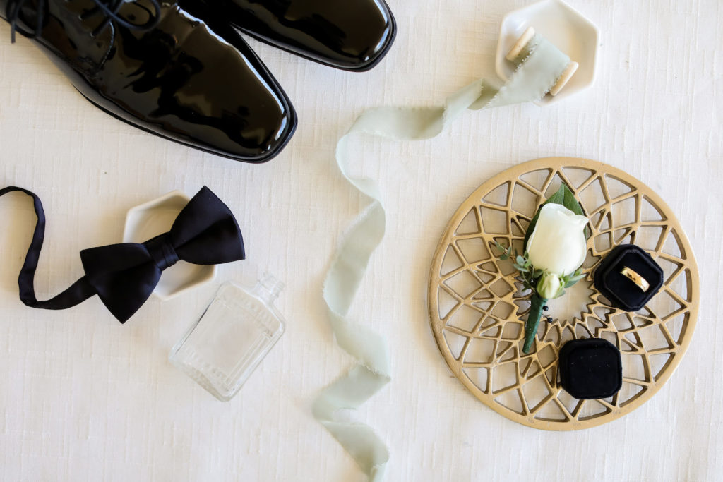 Groom Wedding Accessories, Black Bowtie, Glossy Black Tuxedo Dress Shoes, Bottle of Cologne, Yellow Gold Wedding Ring in Black Velvet Ring Box On Gold Geometric Tray | Tampa Bay Wedding Photographer Lifelong Photography Studio