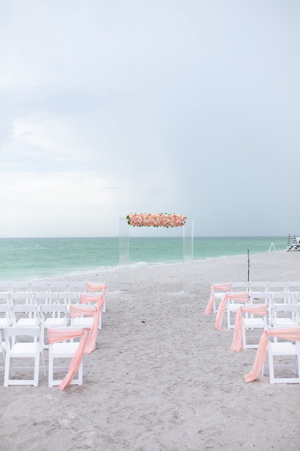 Florida Beach Wedding at Sarasota Wedding Venue The Resort at Longboat Key Club | Ceremony Backdrop with Clear Acrylic Arch and Floral Arrangement of Peach Pink and Coral Roses | White Folding Ceremony Chairs with Peach Sashes down Aisle