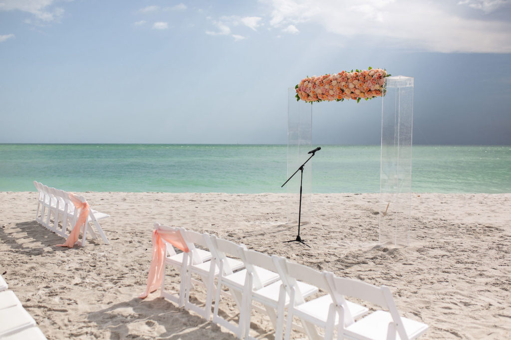 Florida Beach Wedding at Sarasota Wedding Venue The Resort at Longboat Key Club | Ceremony Backdrop with Clear Acrylic Arch and Floral Arrangement of Peach Pink and Coral Roses | White Folding Ceremony Chairs with Peach Sashes down Aisle