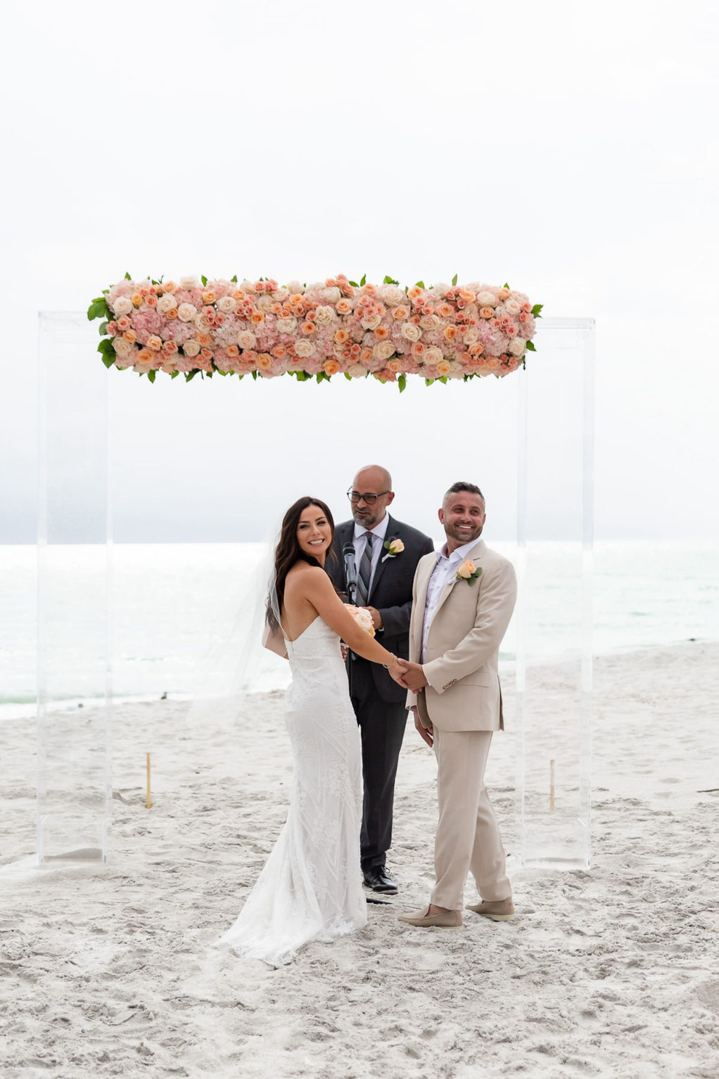 Bride and Groom Exchanging Vows at Florida Beach Wedding at Sarasota Wedding Venue The Resort at Longboat Key Club | Ceremony Backdrop with Clear Acrylic Arch and Floral Arrangement of Peach Pink and Coral Roses | Bride Wearing Strapless Sheath Wedding Gown by Tara Keilly | Groom Wearing Casual Khaki Suit