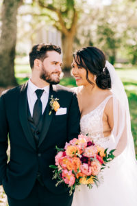 Classic Bride Wearing Plunging V Neckline Lace with Straps and Tulle A-Line Wedding Dress Holding Vibrant Colorful Pink, Orange Roses and Red Floral Bouquet, Groom in Black Tuxedo and Orange Rose Boutonniere | Tampa Bay Wedding Florist Monarch Events and Design