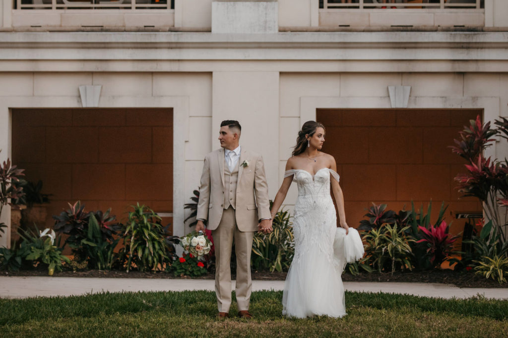 Elegant Bride Wearing Off the Shoulder Mermaid Embellished Sweetheart Neckline Badgley Mischka Wedding Dress Standing Next To and Looking Away from Groom Holding Bridal Floral Bouquet in Tan Suit | Tampa Bay Wedding Dress Shop Truly Forever Bridal