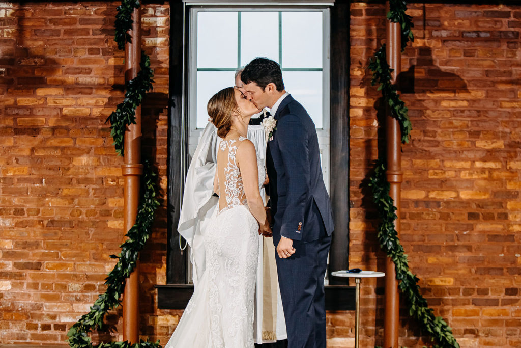 Florida Bride and Groom Kiss During Industrial Inspired Wedding Ceremony, Exposed Brick Warehouse with Ivy and Vine Greenery, Bride Wearing White Lace Wedding Dress with Open Back | Florida Historical Wedding Venue J.C. Newman Cigar Co. in Ybor City