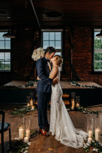 Florida Bride and Groom Kiss During Industrial Inspired Wedding Ceremony, Exposed Brick Warehouse with Ivy and Vine Greenery, Candlelight and flower petals, Bride Wearing White Lace Wedding Dress with Open Back | Florida Historical Wedding Venue J.C. Newman Cigar Co. in Ybor City