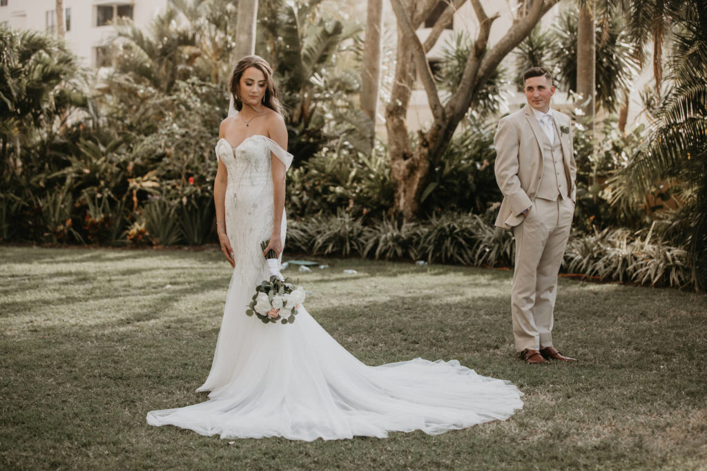 Elegant Bride Wearing Off the Shoulder Mermaid Embellished Sweetheart Neckline Badgley Mischka Wedding Dress Standing Next To and Looking Away from Groom Holding Bridal Floral Bouquet in Tan Suit | Tampa Bay Wedding Dress Shop Truly Forever Bridal