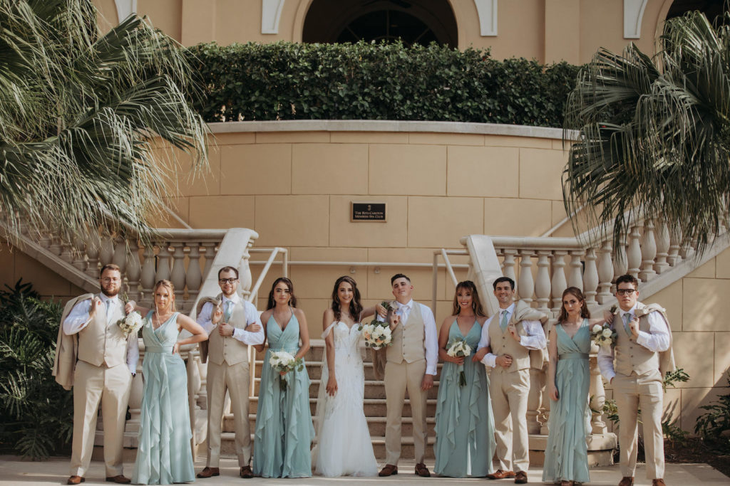 Earthy Elegant Wedding Party, Bridesmaids in Dusty Blue Ruffle Matching Dresses, Groomsmen in Tan Suits and Vests Standing Outside Wedding Venue Ritz Carlton Sarasota | Tampa Bay Wedding Dress Shop Truly Forever Bridal