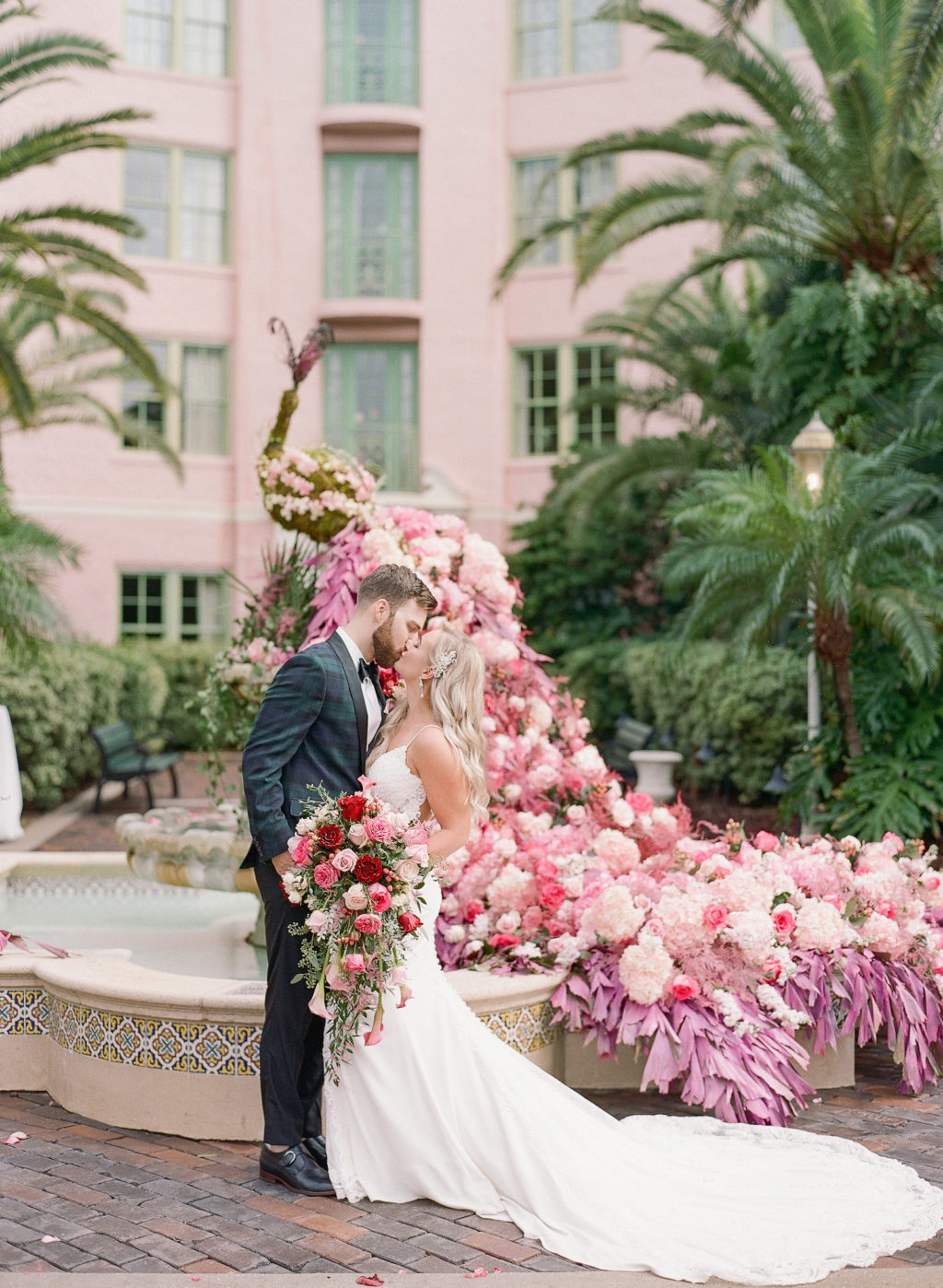 Tampa Bride and Groom Kissing Outside Courtyard by Water Fountain with Floral Peacock Statue with Pink, Red and Purple Floral Tail, Bride Holding Lush Red and Pink Roses with Greenery Floral Bouquet | St. Pete Wedding Venue The Vinoy Renaissance