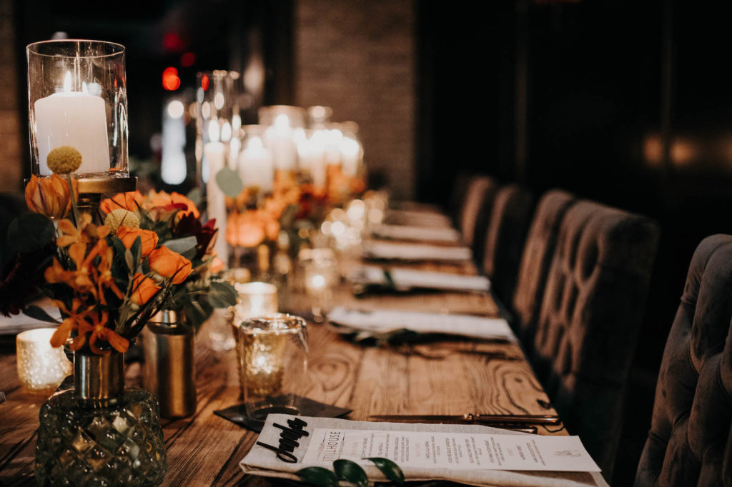 Dark and Moody Wedding Reception Decor, Long Wooden Feasting Table, Black Tufted Chairs, Candlesticks, Burnt Orange Florals | Wedding Venue Urban Stillhouse St. Pete