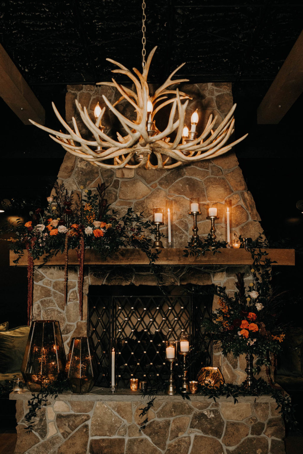 Dark and Moody Wedding Ceremony Decor, Deer Antler Chandelier, Stone Fireplace and Wooden Mantlepiece with Lush Greenery Leaves, Burnt Orang and Blush Pink Roses, Burgundy Hanging Amaranthus, Gold Geometric Speckled Mercury Vases, Candlesticks | Wedding Venue Urban Stillhouse St. Pete