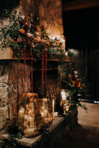 Dark and Moody Wedding Ceremony Decor, Stone Fireplace with Lush Floral Arrangement with Burnt Orange and Blush Pink Roses, Greenery Leaves and Burgundy Hanging Amaranthus, Geometric Speckled Gold Mercury Vases, Candlesticks | Wedding Venue Urban Stillhouse St. Pete