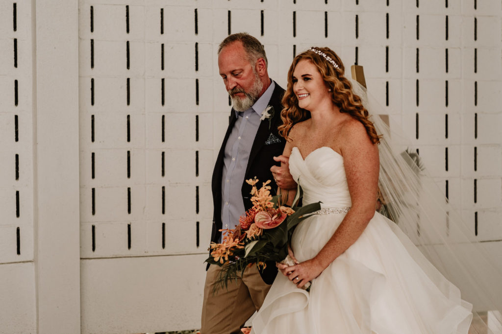 Tropical Elegant Bride Wearing Ballgown Strapless Sweetheart Wedding Dress with Rhinestone Belt Walking Down the Wedding Ceremony Aisle with Dad Holding Palm Fronds, Pink Anthuriums, Pin Cushion and Orange Flower Bouquet | Tampa Bay Wedding Florist Iza's Flowers