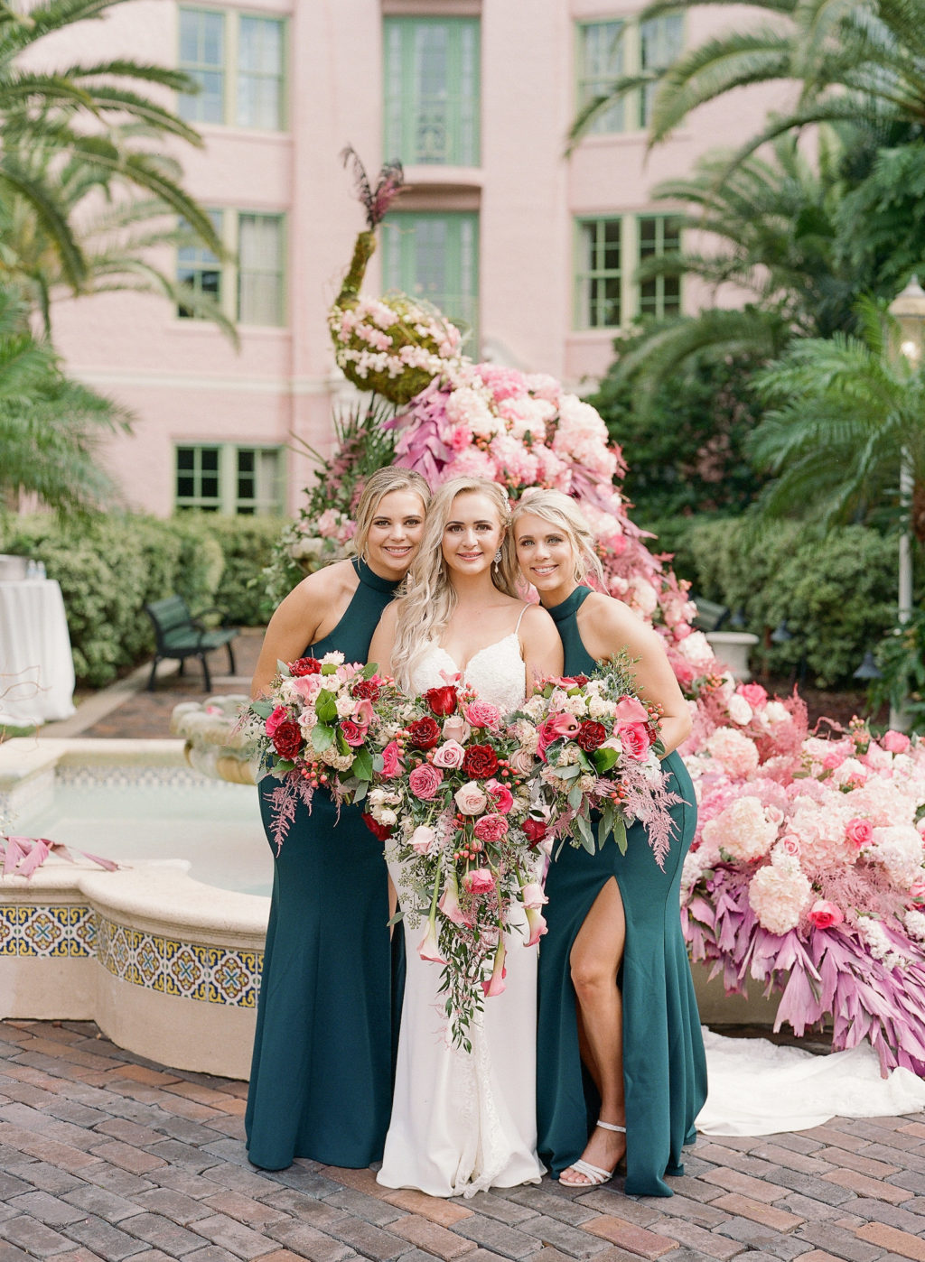 Elegant Bride with Bridesmaids in Emerald Green Dresses Holding Red, Pink and White Roses with Greenery Floral Bouquets Standing in Front of Courtyard Water Fountain with Floral Peacock Statue with Pink, White and Purple Lush Floral Tail | St. Pete Wedding Venue The Vinoy Renaissance | Tampa Bay Wedding Hair and Makeup Femme Akoi Beauty Studio