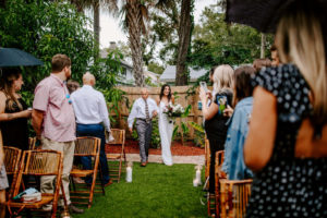 Tampa Bay Bride and Father Walk Down the Aisle in Private Backyard Wedding Ceremony, Holding Lush Tropical Bouquet, Jungle Inspired Decor with Bamboo Folding Chair Rentals | Florida Wedding Planner Parties A'La Carte