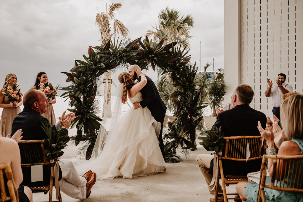 Florida Bride and Groom Exchanging Wedding Vows Under Tropical Circular Palm Fronds and Leaves Circular Wedding Ceremony Arch | Waterfront Wedding Venue Hilton Clearwater Beach | Tampa Bay Wedding Florist Iza's Flowers