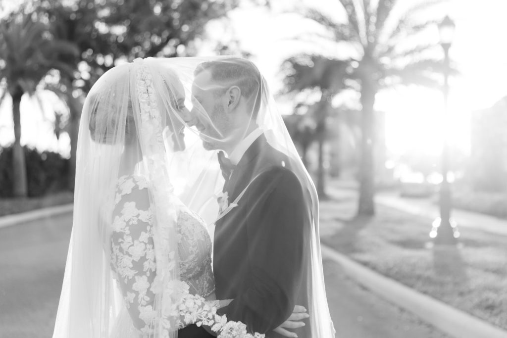 Romantic Florida Bride and Groom Kiss Under Veil with Sunset and Palm Trees in the Backdrop, Bride Wearing Adam Zhoar Wedding Dress with Illusion Lace Sleeves, Galia Lahav Veil, Groom Wearing Classic Black Tie Giorgio Armani Tux | Florida Bridal BoutiqueRomantic Florida Bride and Groom Kiss Under Veil with Sunset and Palm Trees in the Backdrop, Bride Wearing Adam Zhoar Wedding Dress with Illusion Lace Sleeves, Galia Lahav Veil, Groom Wearing Classic Black Tie Giorgio Armani Tux | Florida Bridal Boutique Isabel O'Neil Bridal Collection
