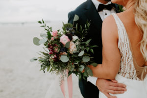 Florida Bride in Fitted Lace and Illusion Plunging V Open Back Wedding Dress and Full Length Veil Holding Wild Greenery Eucalyptus with Pink and Purple Floral Bouquet, Groom in Emerald Green Tuxedo Jacket and Bowtie | St. Pete Wedding Venue Postcard Inn on the Beach