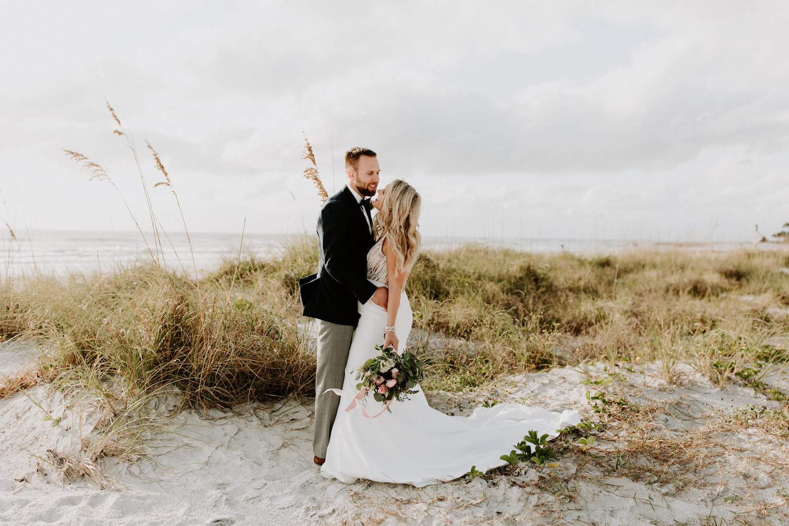 Florida Bride in Fitted Lace and Illusion Plunging V Open Back Wedding Dress and Full Length Veil Holding Wild Greenery Eucalyptus with Pink and Purple Floral Bouquet, Groom in Emerald Green Tuxedo Jacket and Bowtie | St. Pete Wedding Venue Postcard Inn on the Beach