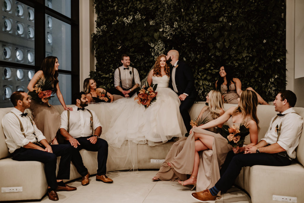 Florida Wedding Party Portrait, Bride in Ballgown Wedding Dress Holding Tropical Floral Bouquet, Groom Kissing Bride, Bridesmaids Wearing Beige Dresses, Groomsmen in Suspenders and Bow Ties Sitting on Curved Sofa | Tampa Bay Wedding Florist Iza's Flowers | Wedding Venue Hilton Clearwater Beach