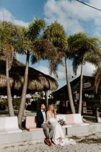 Florida Bride and Groom Sitting on Bench of Sandy Beach | St. Pete Wedding Venue Postcard Inn on the Beach