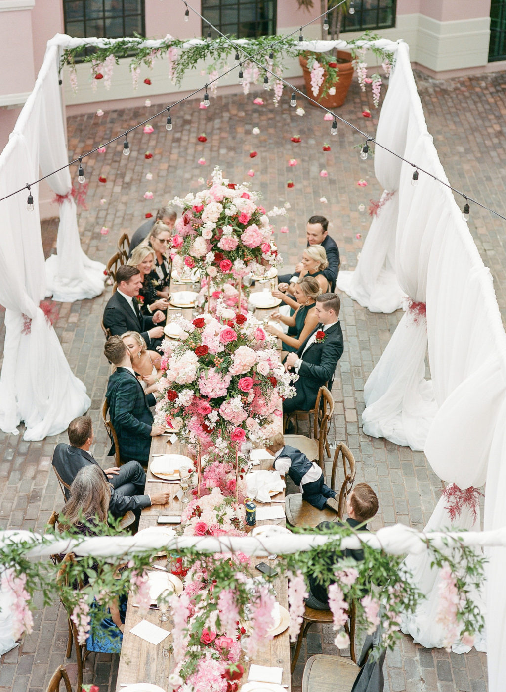 Wedding Guests Sitting At Elegant Garden Themed Wedding Reception, Long Wooden Feasting Table in Outdoor Hotel Courtyard, White Drapery with Hanging Flowers and String Lights, Lush Pink, Red Roses and White Flower Centerpieces and Table Runner