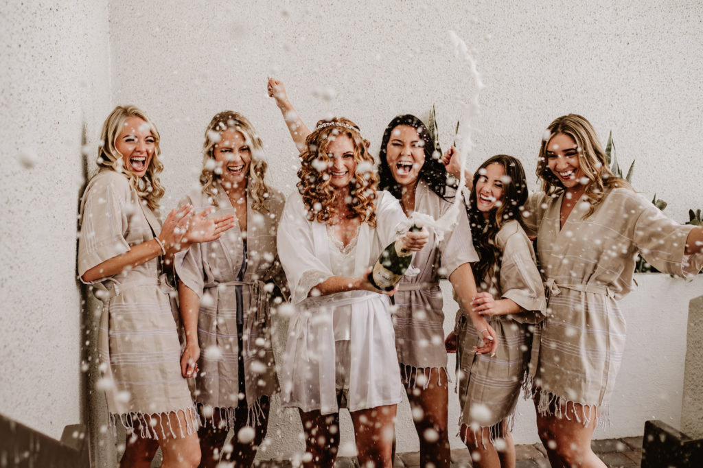 Bride and Bridesmaids Popping Bottle of Champagne in Oatmeal Neutral Color Matching Robes Getting Wedding Ready